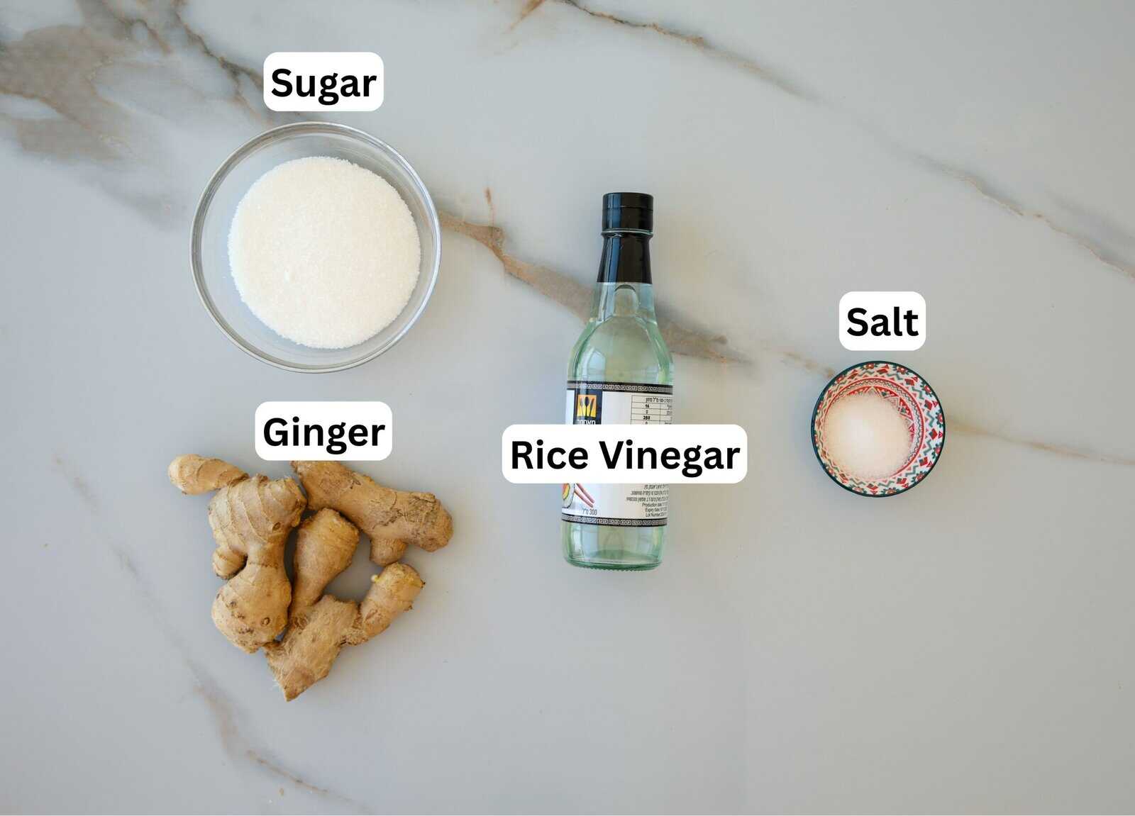 A bowl of sugar, a piece of ginger, a bottle of rice vinegar, and a small bowl of salt are arranged on a marble surface, each labeled with its name.