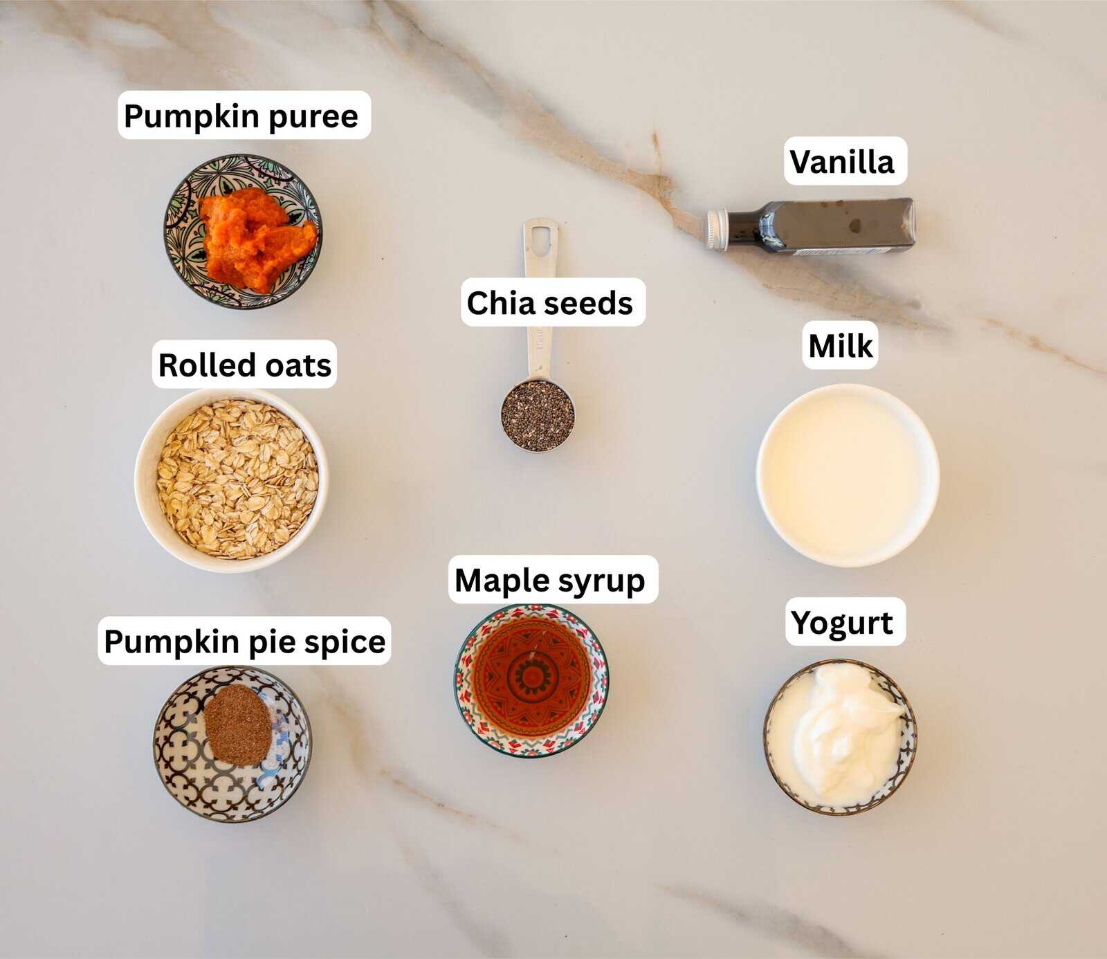 Seven ingredients for a pumpkin recipe are arranged on a marble surface: pumpkin puree, rolled oats, pumpkin pie spice, chia seeds, maple syrup, milk, yogurt, and a bottle of vanilla, each labeled.