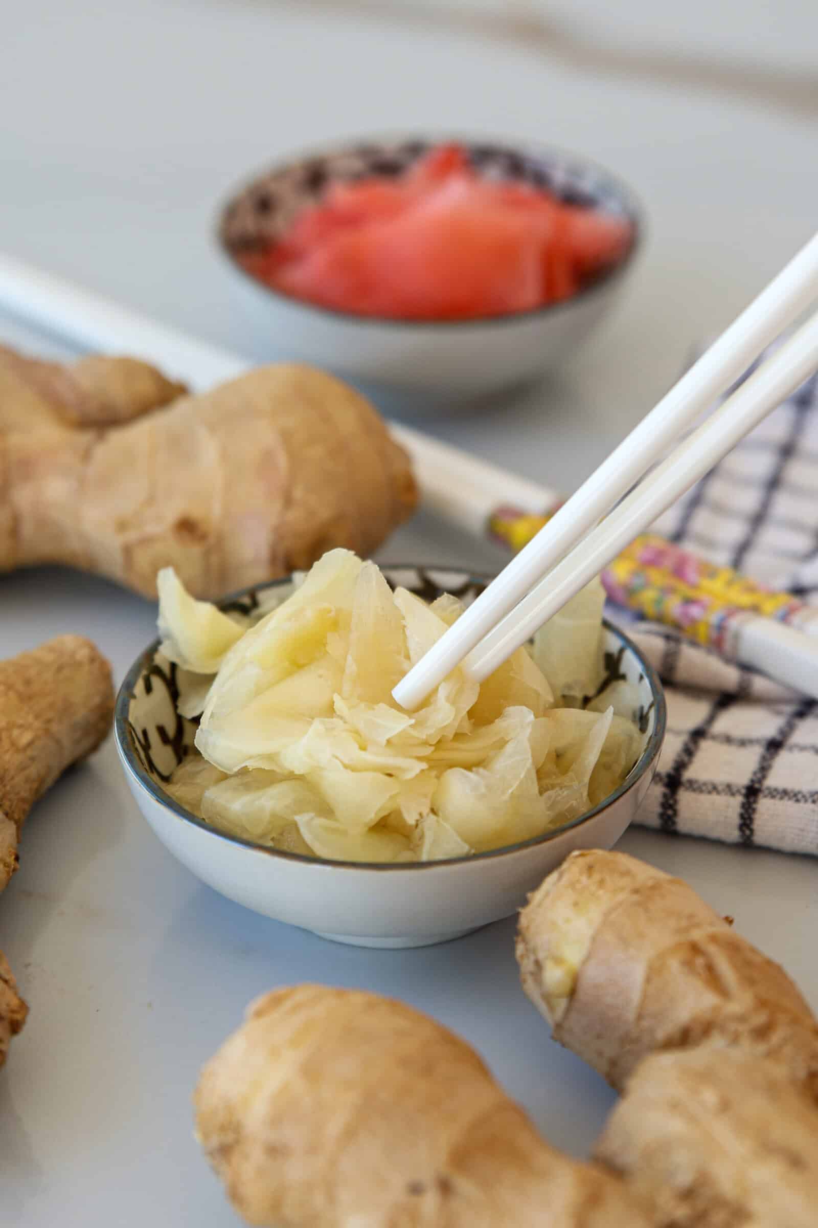 A small bowl filled with pickled ginger being picked up with chopsticks, surrounded by fresh ginger roots. Another bowl with pink pickled ginger is blurred in the background. A striped napkin is also visible.