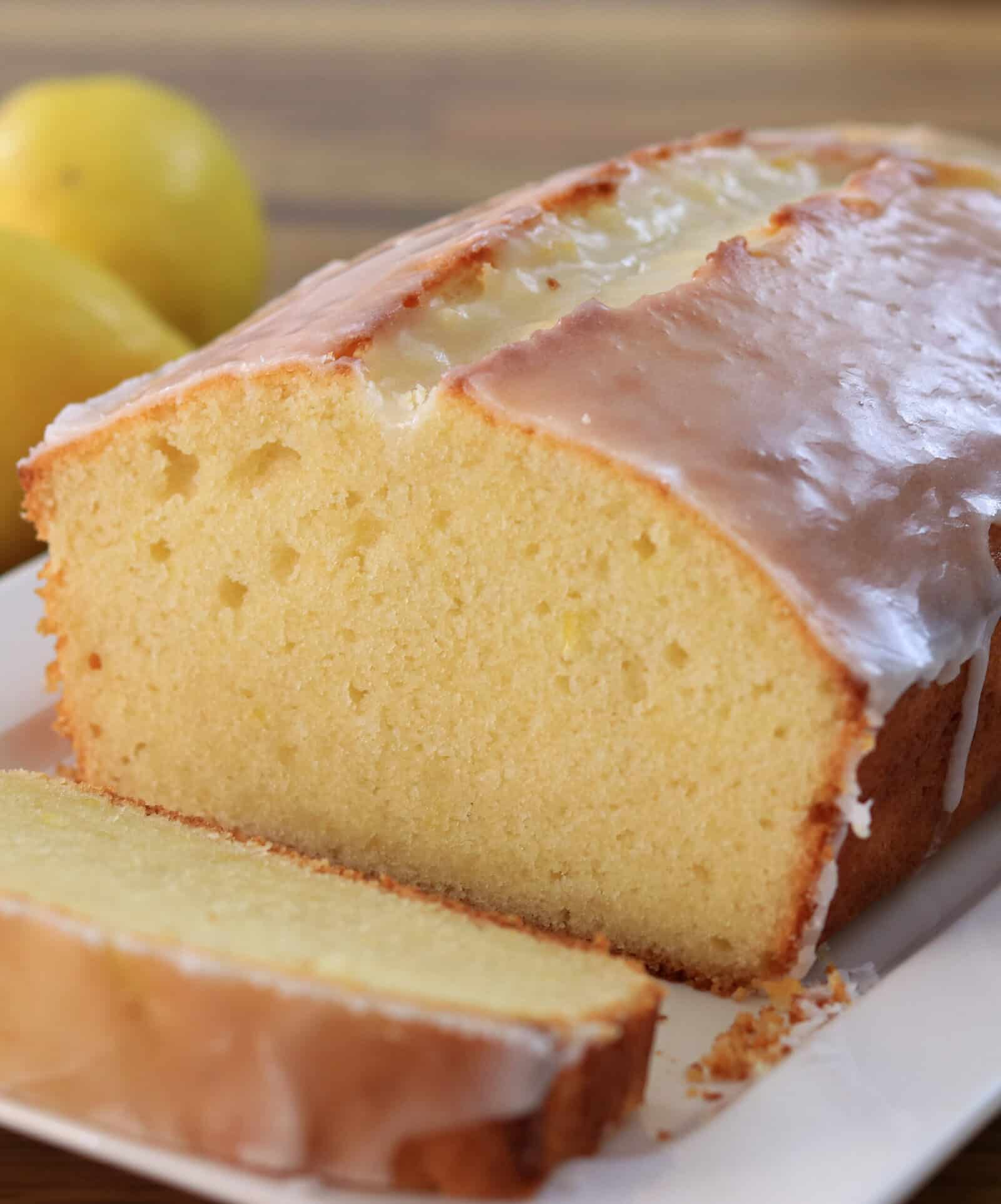 A close-up of a loaf of lemon pound cake with a glossy glaze on top, partially sliced on a white plate. Two whole lemons are visible in the background.