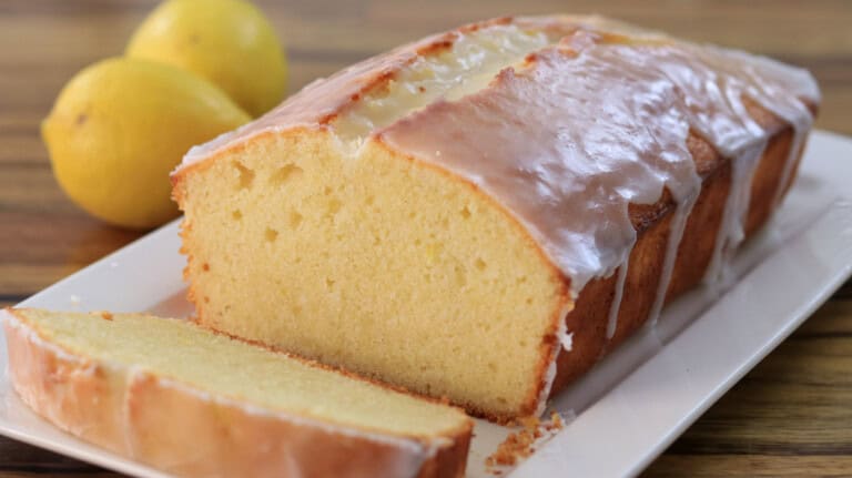 A loaf of glazed lemon pound cake on a white rectangular plate, with one slice cut. Two whole lemons are in the background on a wooden surface.
