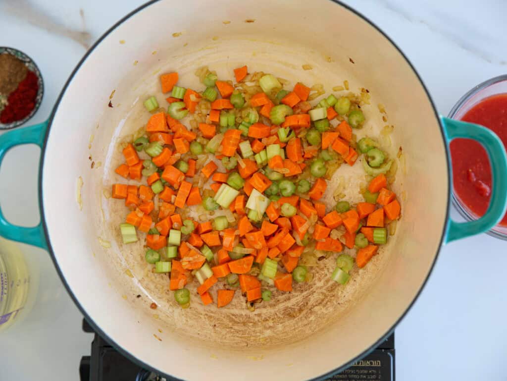 Diced carrots, celery, and onions are sautéing in a large, light-colored pot on a stovetop, with a bowl of tomato sauce and spices visible nearby.