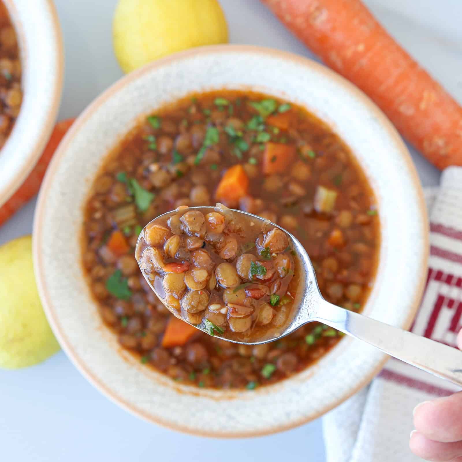 A spoonful of lentil soup is held above a bowl filled with the same soup, garnished with herbs and diced carrots. A carrot, lemons, and a red-striped towel are visible in the background.
