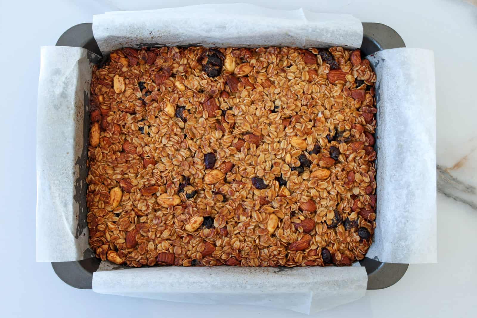 A tray of freshly baked granola bars with visible oats, nuts, and dried fruit, lined with white parchment paper and set on a white marble surface.