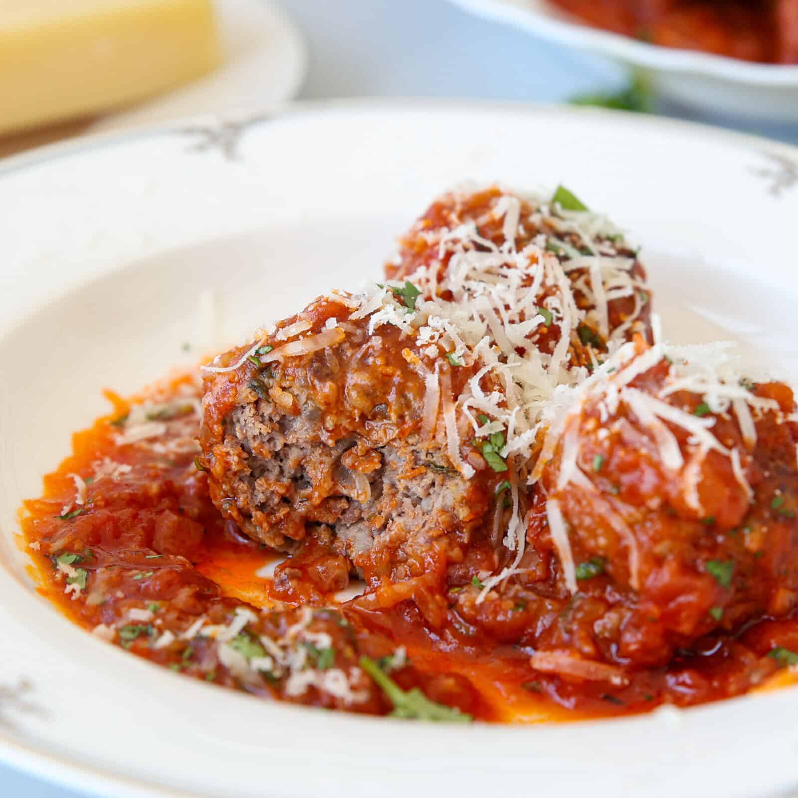 Two meatballs topped with grated cheese sit in a rich tomato sauce on a white plate, garnished with chopped herbs.