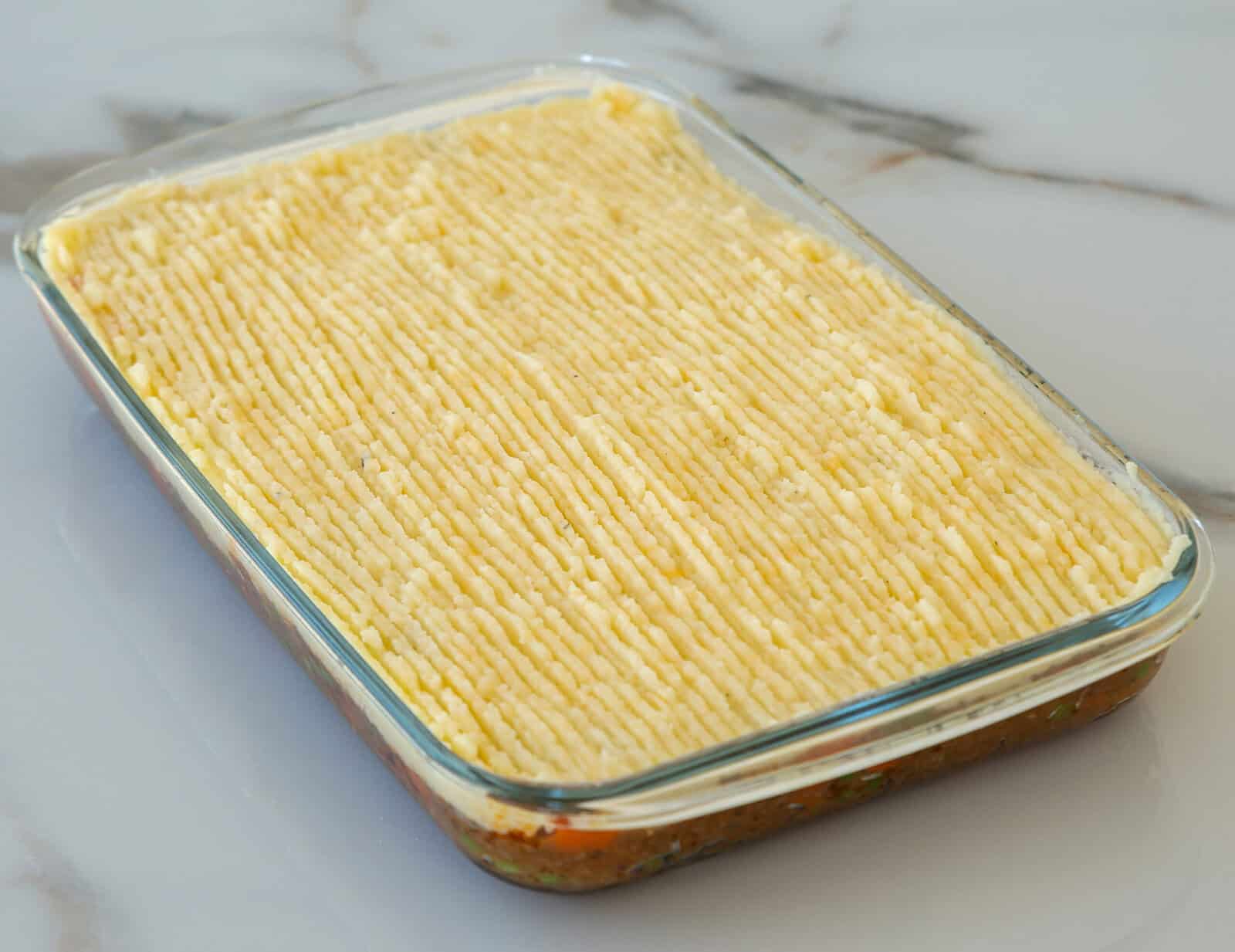 A glass baking dish filled with shepherd’s pie, topped with evenly textured piped mashed potatoes, sits on a marble surface. The filling is visible along the sides of the dish.