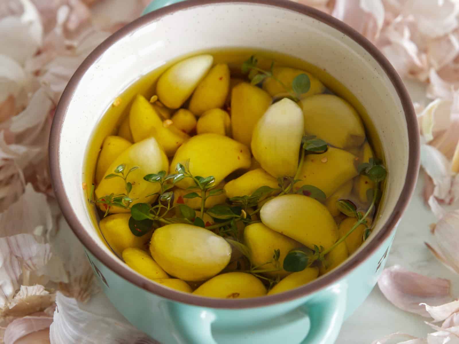 A turquoise pot filled with garlic cloves, fresh herbs, and olive oil, surrounded by loose garlic skins.