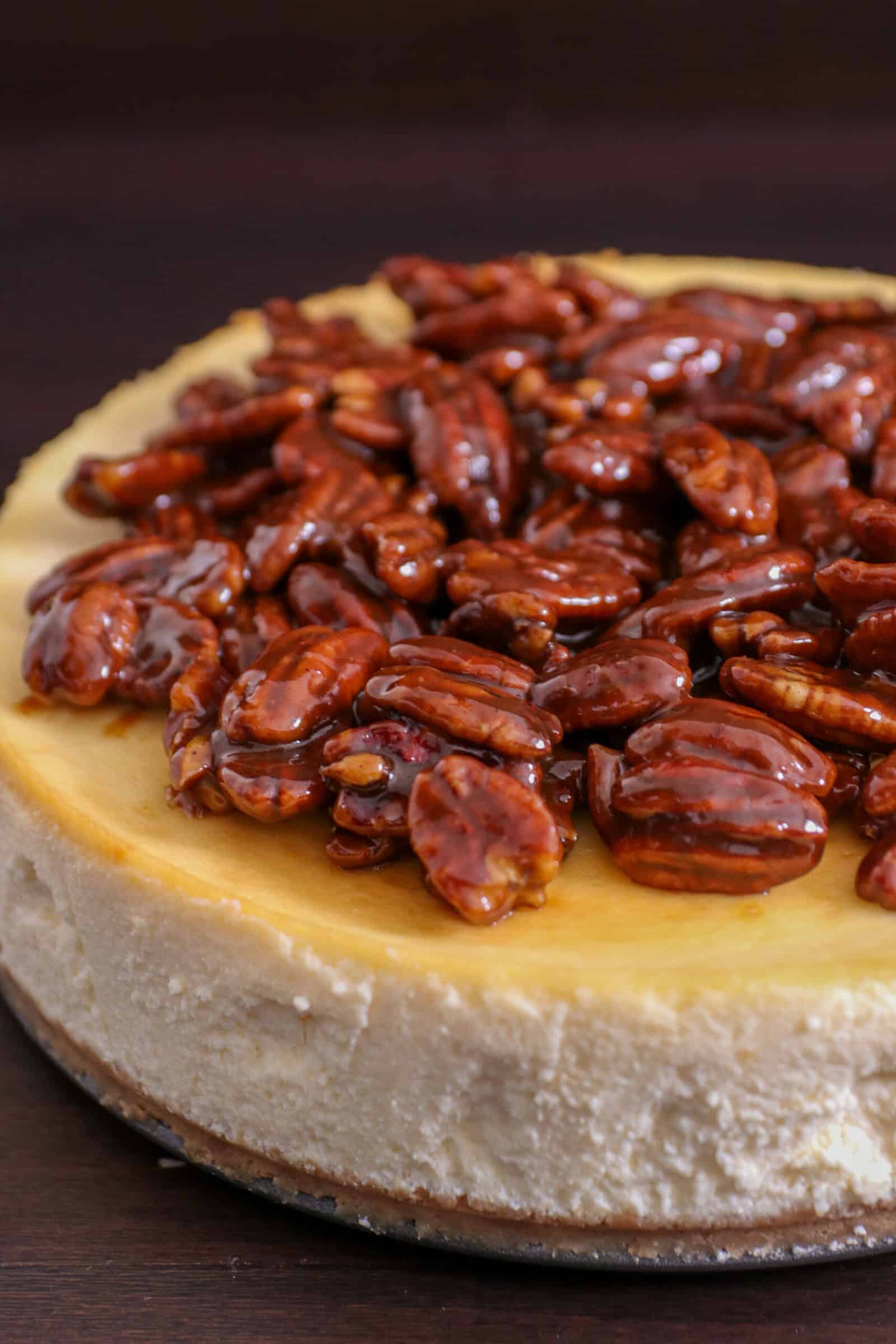 A cheesecake topped with glossy, caramelized pecans sits on a dark surface, showcasing its creamy texture and nutty topping.