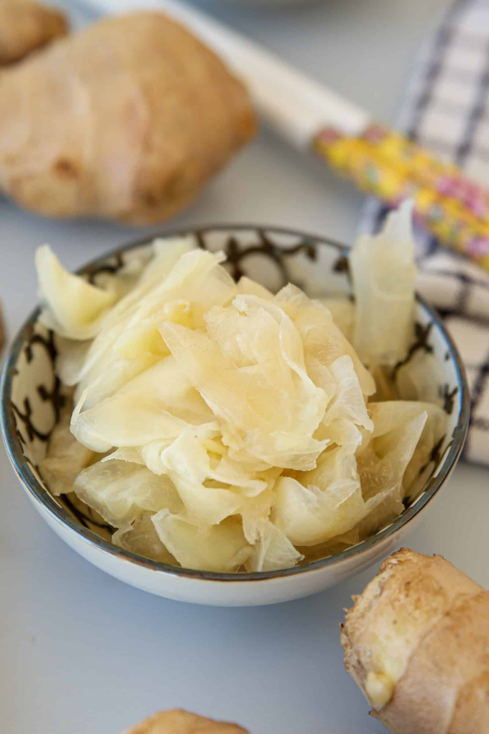 A bowl filled with thin, pale slices of pickled ginger sits on a table with fresh ginger roots and a patterned cloth nearby.