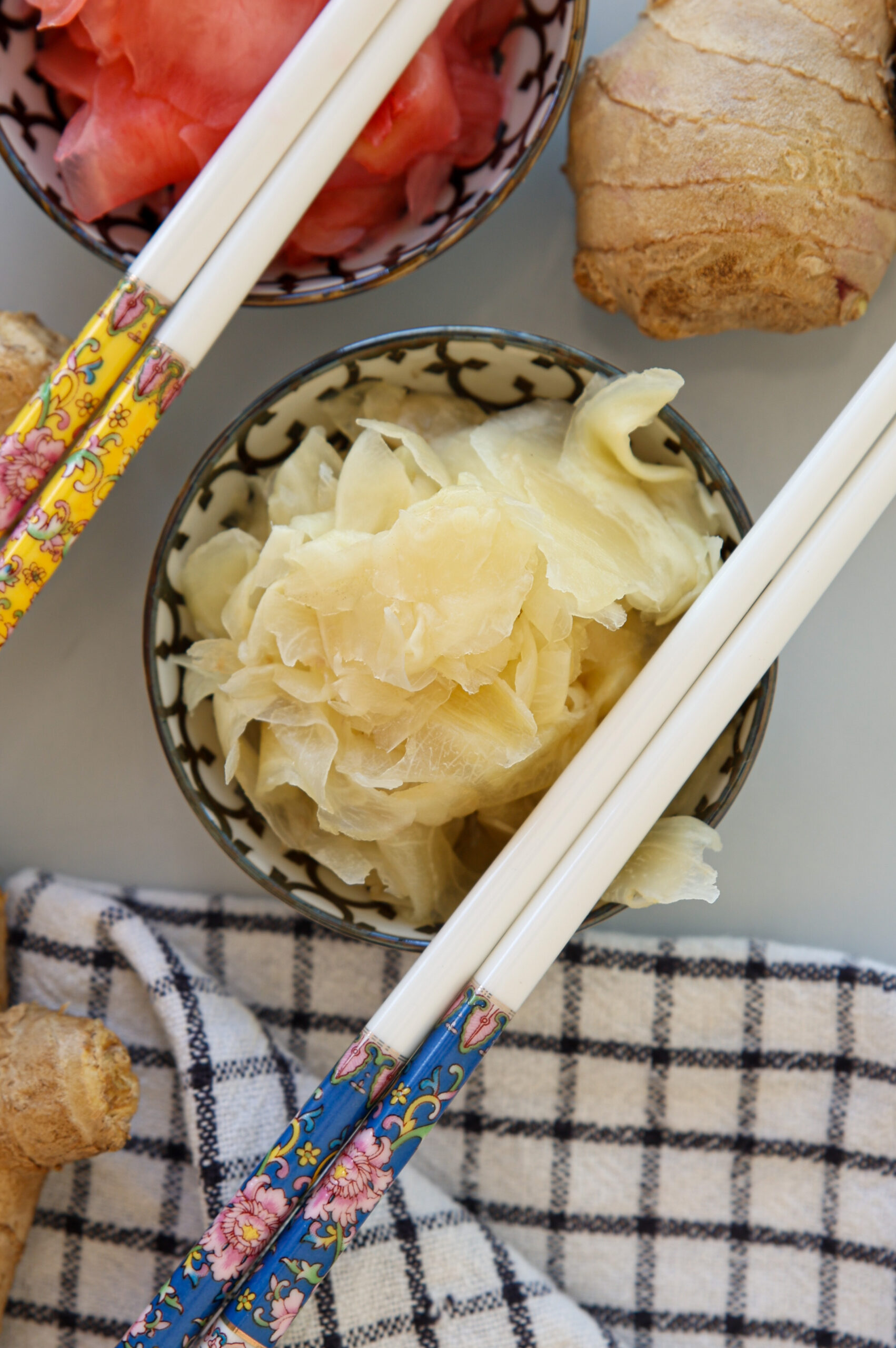A bowl of sliced pickled ginger is placed on a checkered cloth, surrounded by colorful chopsticks, fresh ginger root, and another bowl of pink pickled ginger.