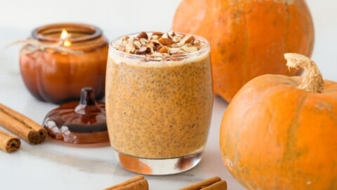 Healthy Pumpkin Chia Pudding Recipe A glass of pumpkin chia pudding topped with chopped nuts sits on a white surface, surrounded by pumpkins, cinnamon sticks, and a lit pumpkin-shaped candle.