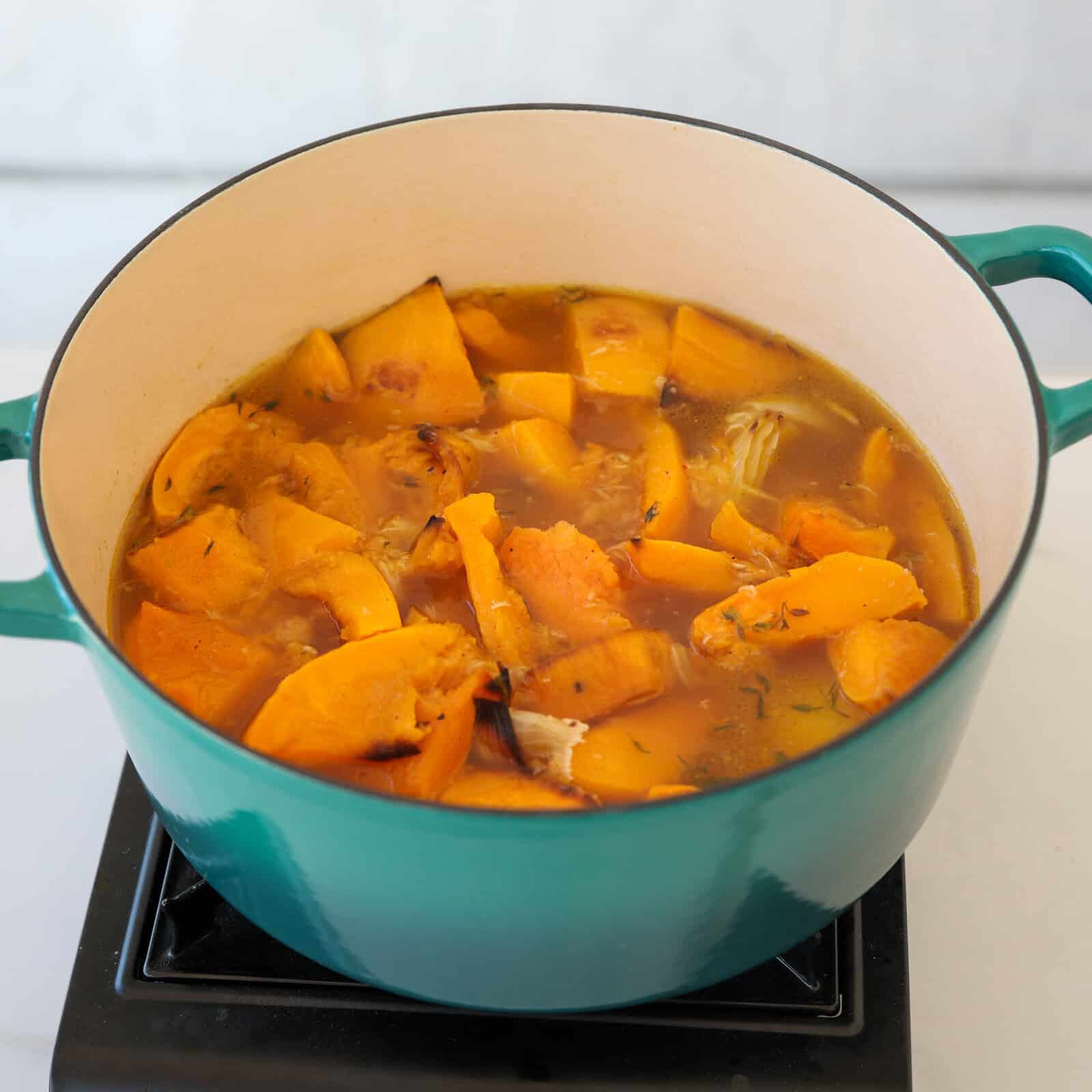 Chunks of orange pumpkin simmer in a light broth inside a large, green, enameled pot on a stovetop. The soup appears to be cooking, with steam rising gently.
