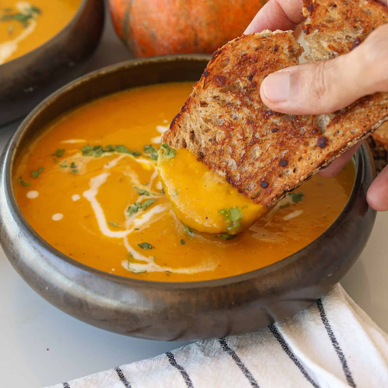 A hand dips a slice of toasted bread into a bowl of creamy orange soup, garnished with a swirl of cream and chopped herbs. A striped cloth napkin and a pumpkin are visible in the background.