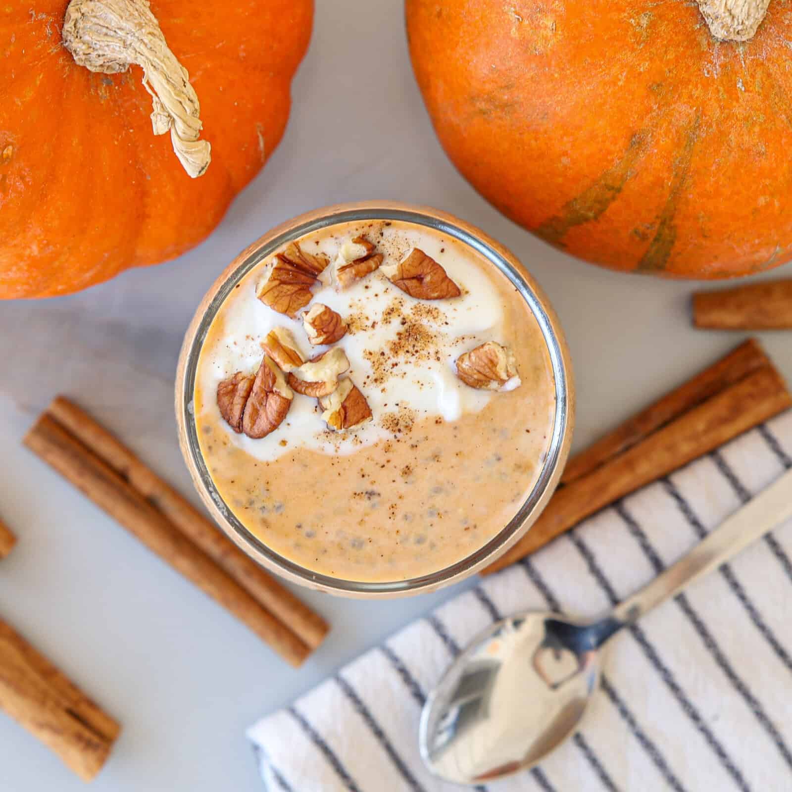A glass of pumpkin pie overnight oats topped with whipped cream, chopped pecans, and a sprinkle of spice. Two pumpkins, cinnamon sticks, a striped napkin, and a spoon surround the glass on a light surface.
