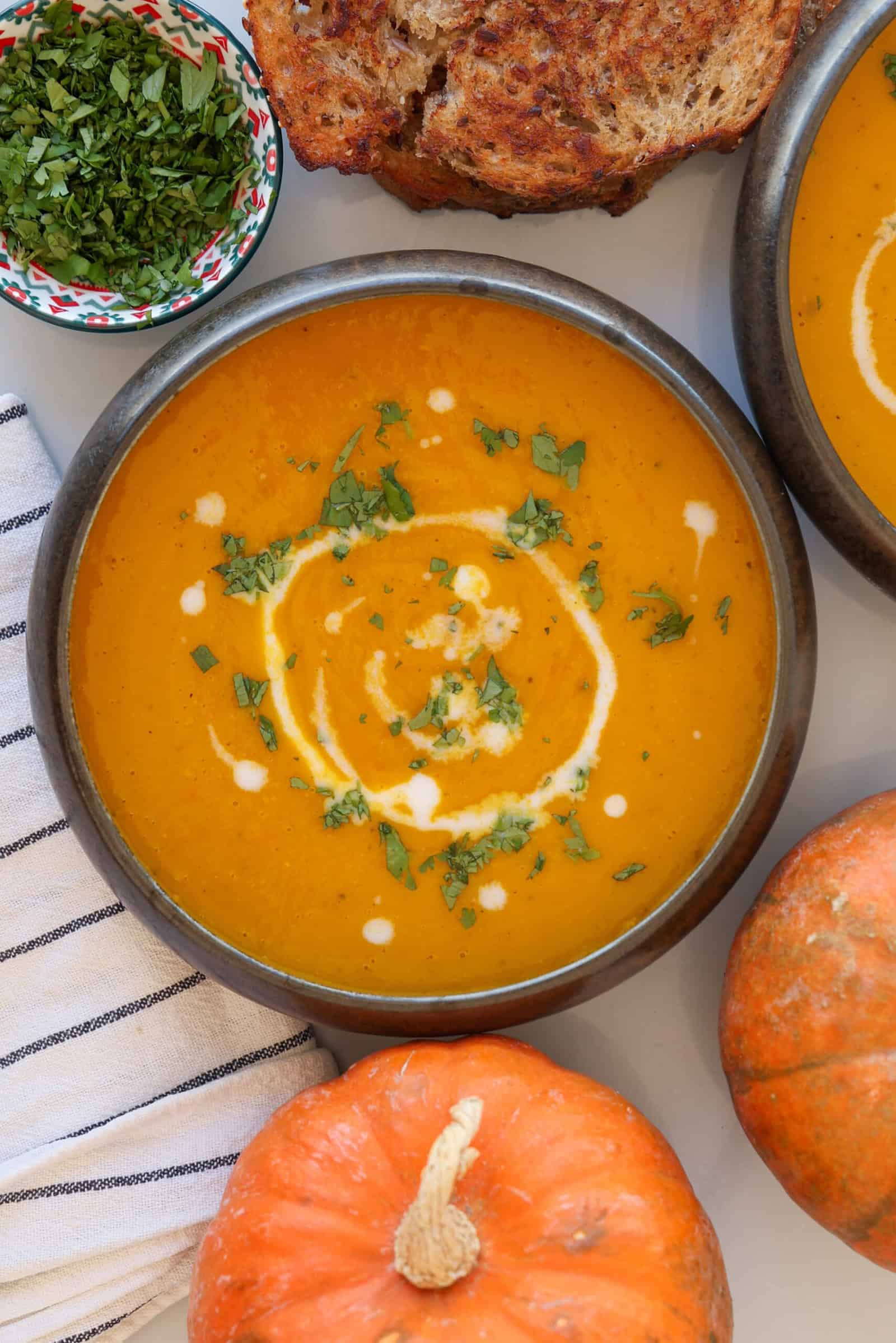 A bowl of pumpkin soup garnished with cream and chopped herbs, next to sliced bread, a small bowl of fresh herbs, a striped towel, and two small pumpkins.
