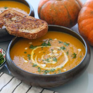 A bowl of creamy orange pumpkin soup garnished with chopped herbs and a swirl of cream, served with a slice of toasted bread. Whole pumpkins and a striped napkin are nearby on the table.