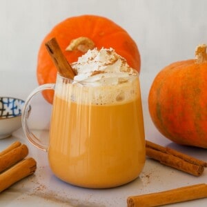 A glass mug of pumpkin spice latte topped with whipped cream, cinnamon, and a cinnamon stick, surrounded by pumpkins and loose cinnamon sticks on a white surface.