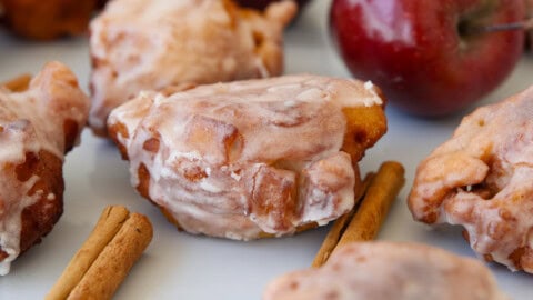 Close-up of glazed apple fritters with pieces of apple inside, surrounded by whole cinnamon sticks and a red apple in the background.