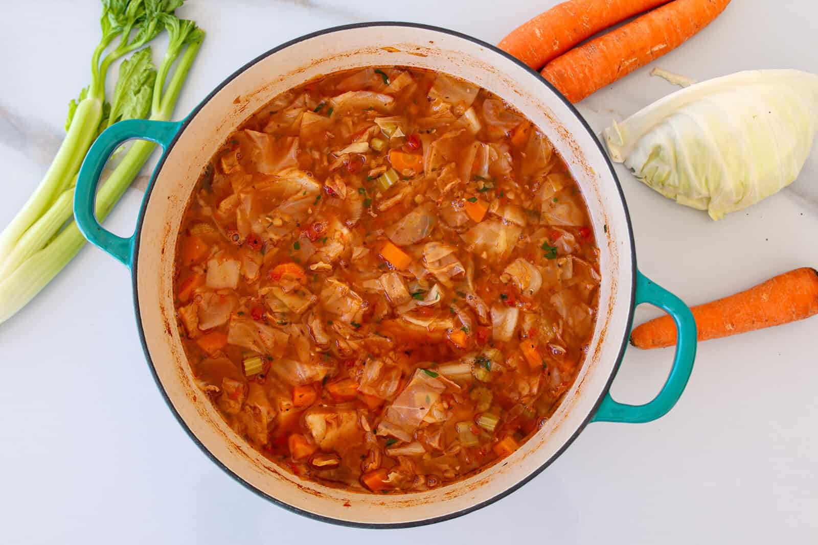 A pot of homemade vegan cabbage soup with visible chunks of cabbage, carrots, and celery, surrounded by fresh carrots, cabbage, and celery on a white countertop.