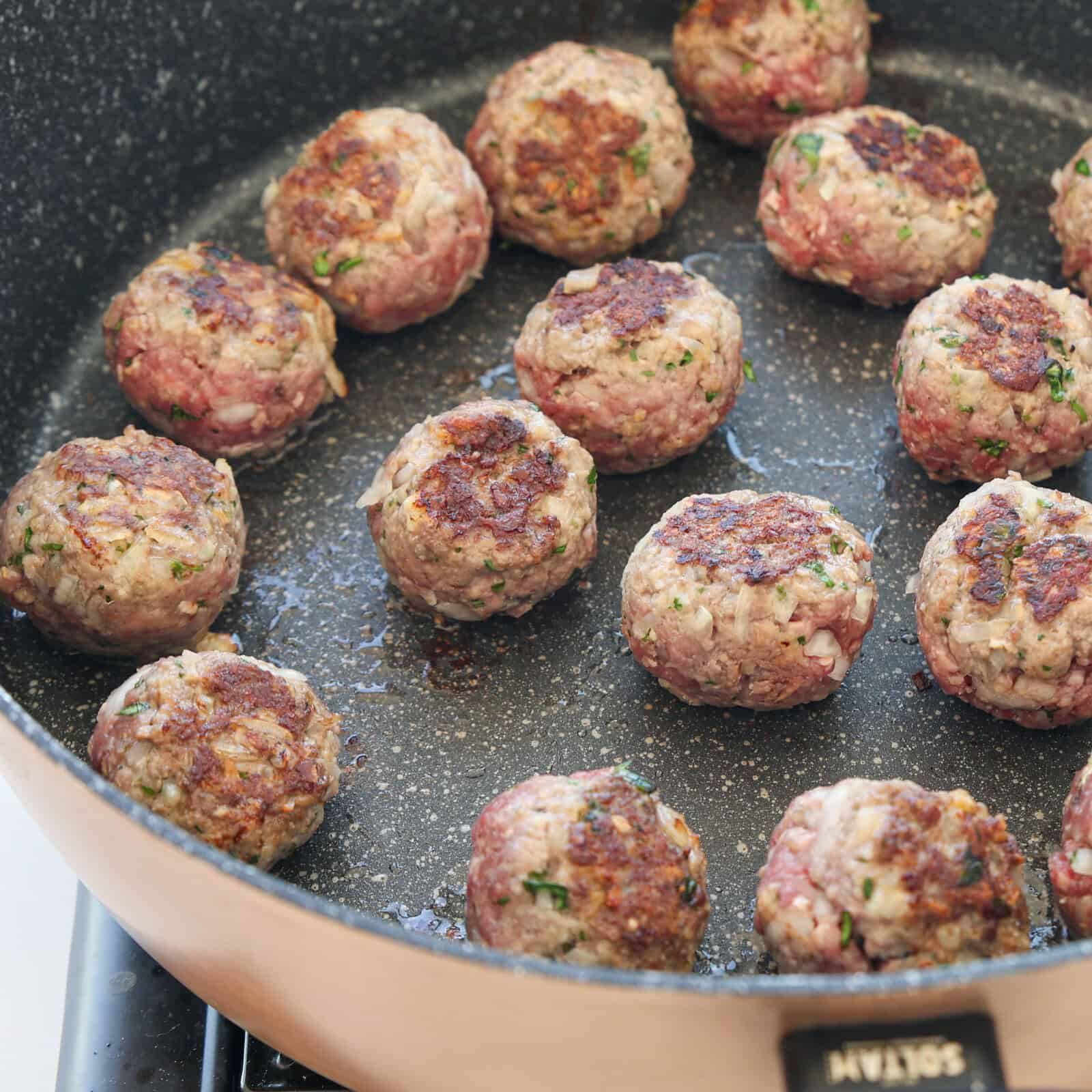 Meatballs are being browned in a nonstick frying pan on a stovetop. The meatballs have a golden-brown crust and are spaced evenly in the pan.