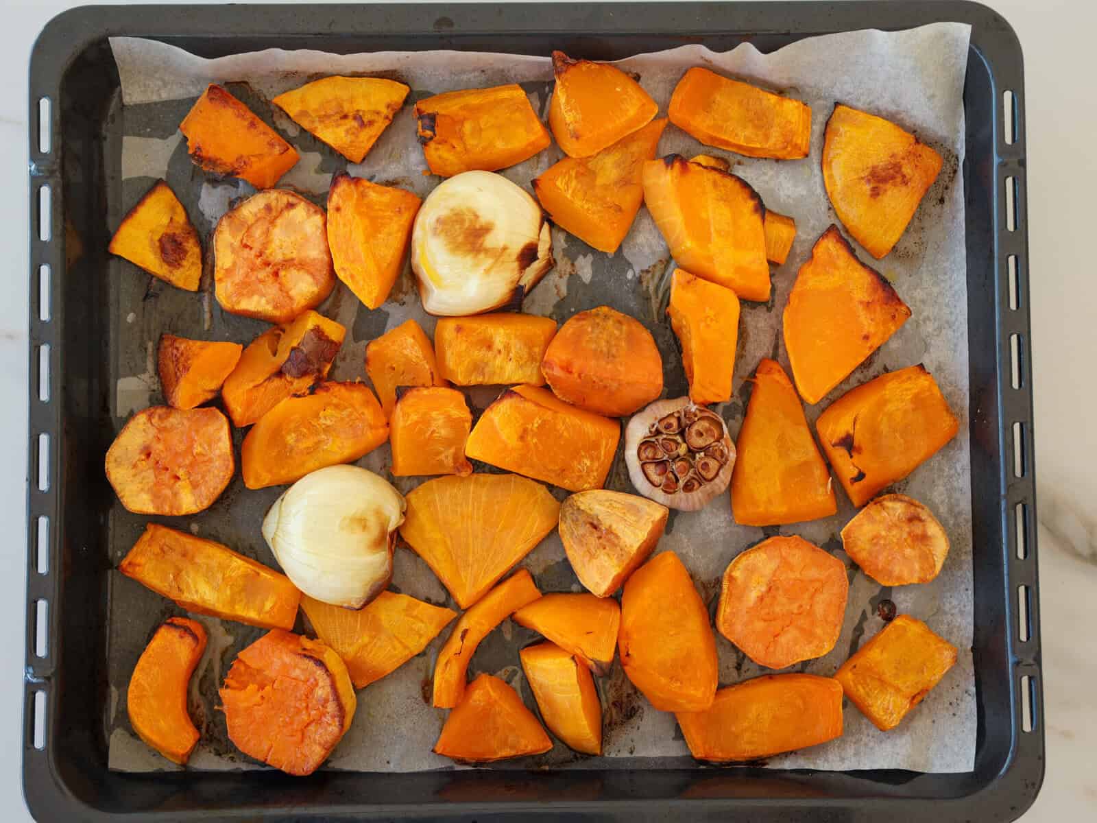 A baking tray lined with parchment paper holds roasted orange vegetables, including pumpkin and sweet potato chunks, along with roasted halved onions and a halved garlic bulb.