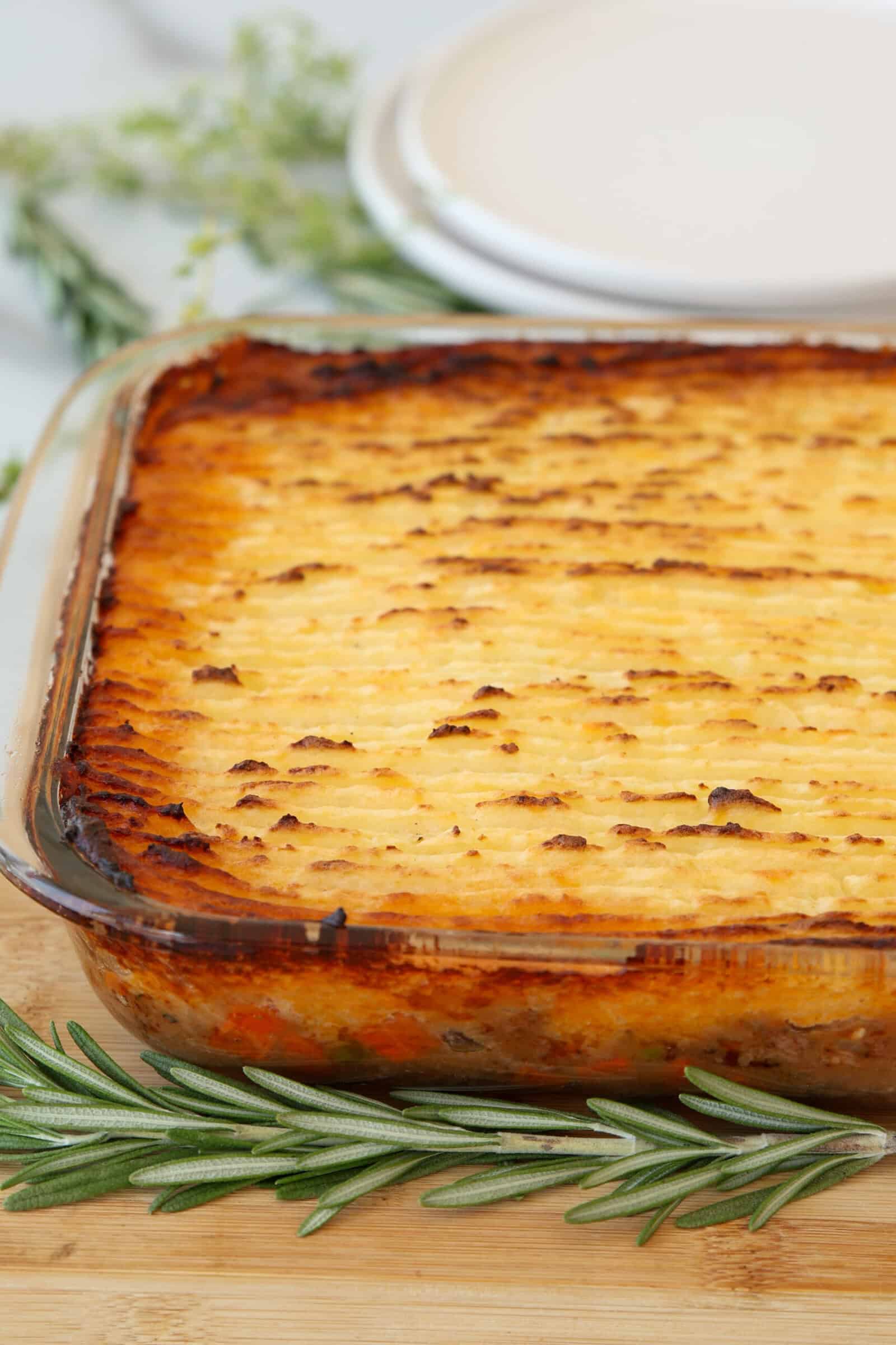 A golden-brown shepherd’s pie in a glass baking dish sits on a wooden surface with sprigs of fresh rosemary nearby and plates in the blurred background.