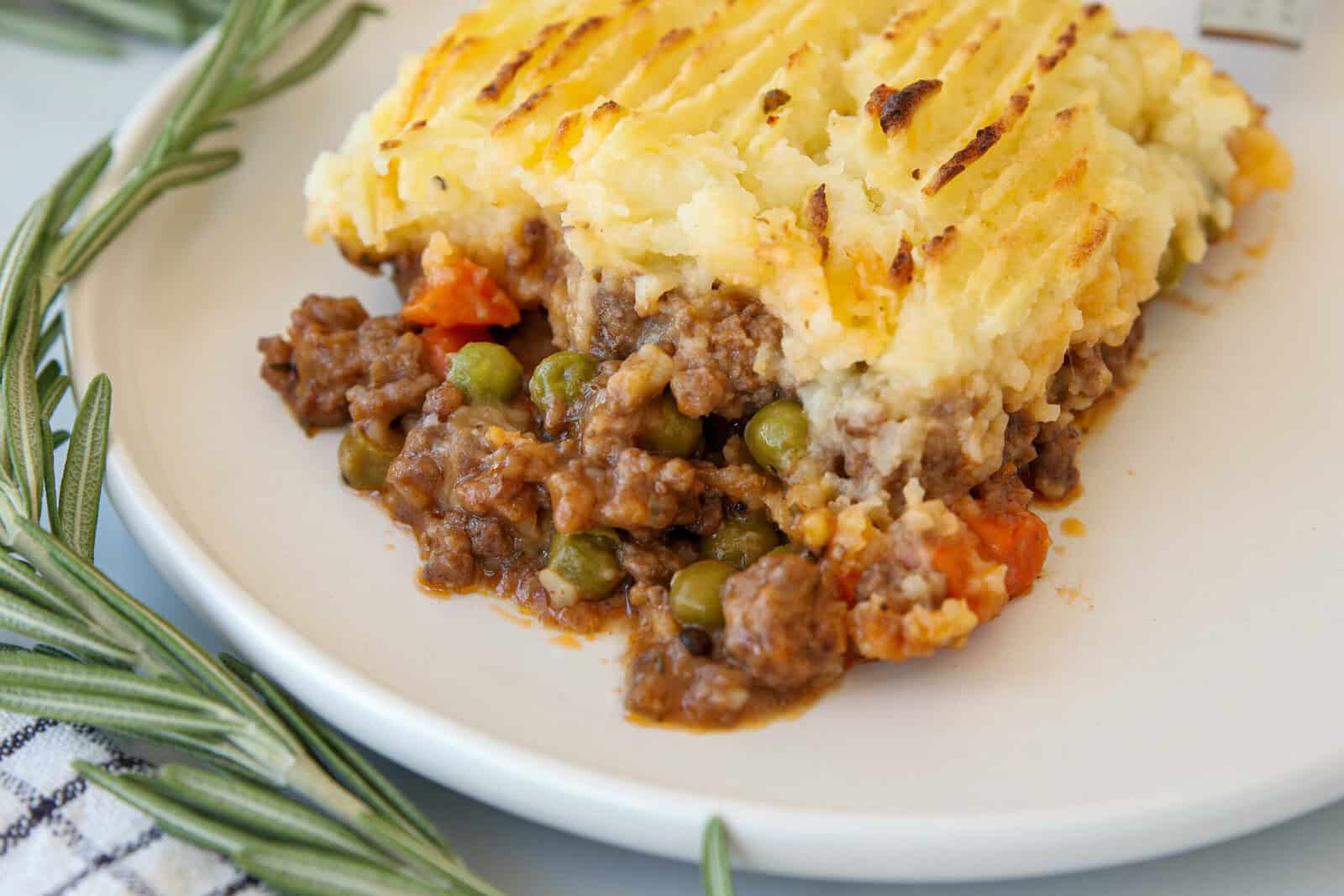 A serving of shepherd's pie on a white plate, featuring a golden, crispy mashed potato topping over ground meat, peas, and carrots. Fresh rosemary sprigs and a striped cloth are nearby.