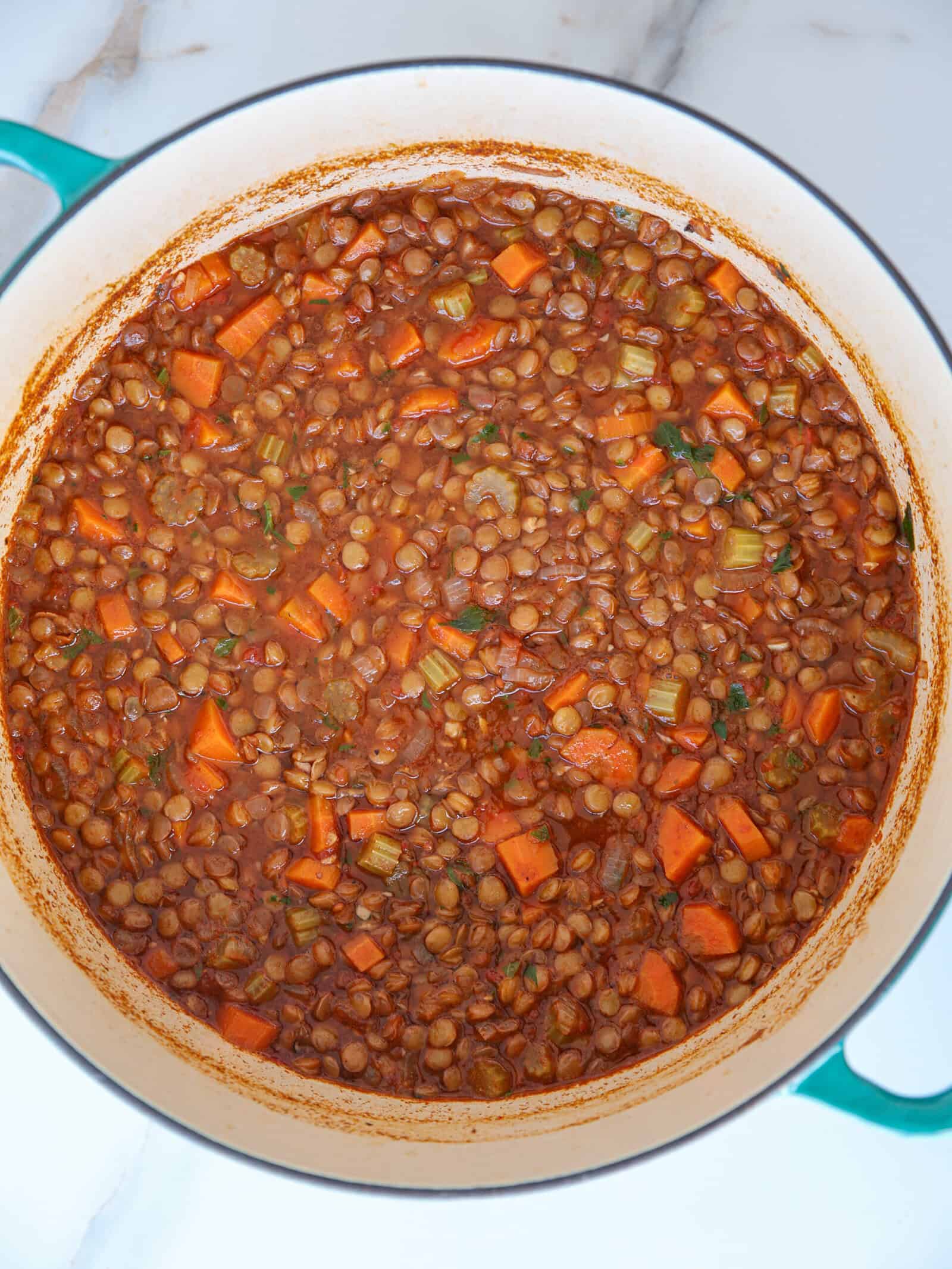 A pot filled with hearty lentil stew, containing lentils, diced carrots, celery, and herbs in a tomato-based broth, sits on a white marble surface.