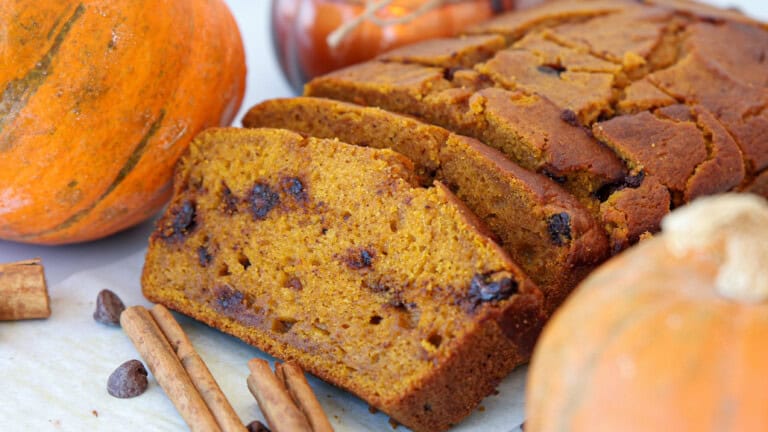 A loaf of pumpkin bread with chocolate chips, sliced to reveal its moist texture, sits on a table surrounded by pumpkins, cinnamon sticks, and chocolate chips.