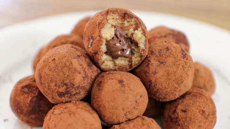 A plate of tiramisu truffles coated in cocoa powder, with one truffle on top bitten open to reveal a creamy chocolate filling inside.