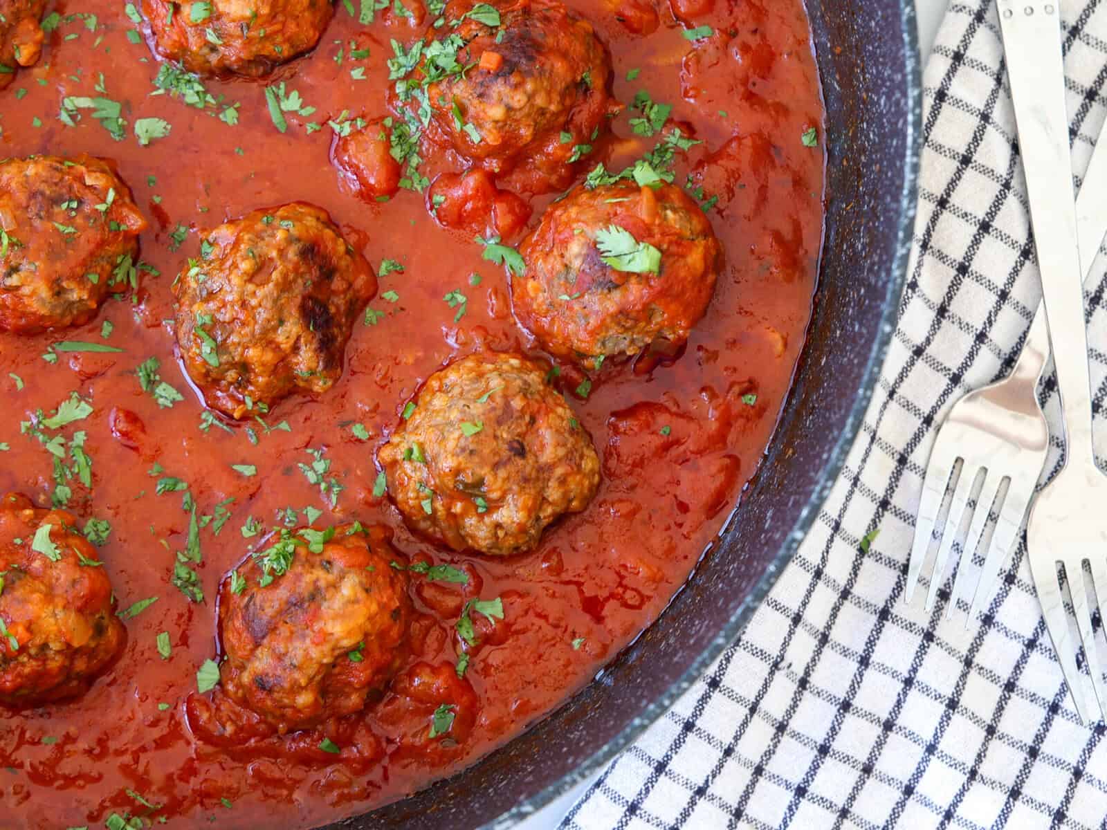 Meatballs simmering in a pan of tomato sauce, garnished with chopped herbs. The pan rests on a checkered cloth, with two metal forks placed on the side.
