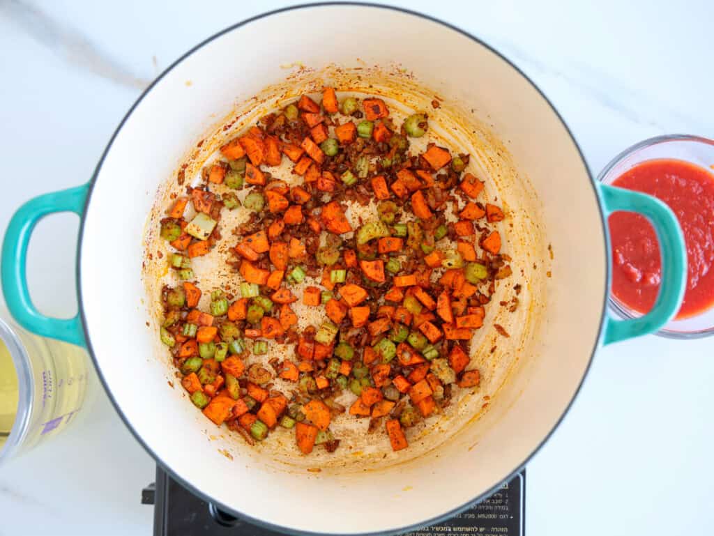 Chopped carrots and celery are being sautéed with seasonings in a large, round, teal-handled pot on a stovetop; a bowl of tomato sauce and a container of oil are nearby.