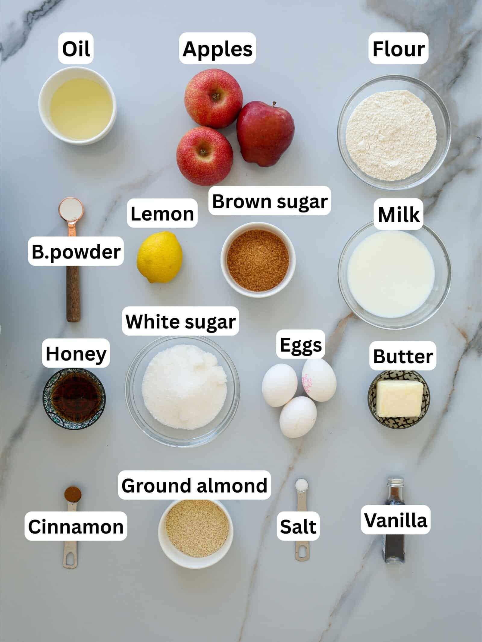 Various baking ingredients labeled on a marble surface, including oil, apples, flour, lemon, brown sugar, milk, baking powder, white sugar, eggs, butter, honey, ground almond, cinnamon, salt, and vanilla.
