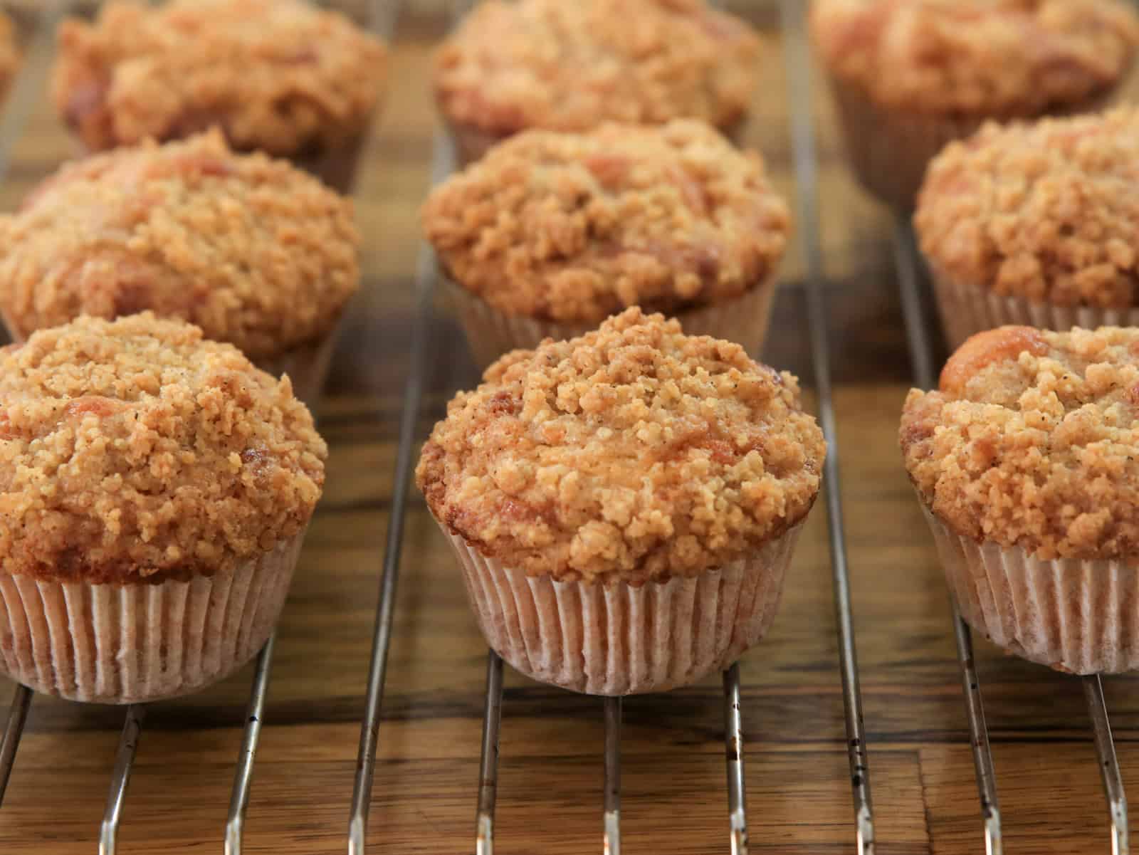 A close-up of freshly baked apple muffins with crumb topping, arranged in rows on a metal cooling rack over a wooden surface.