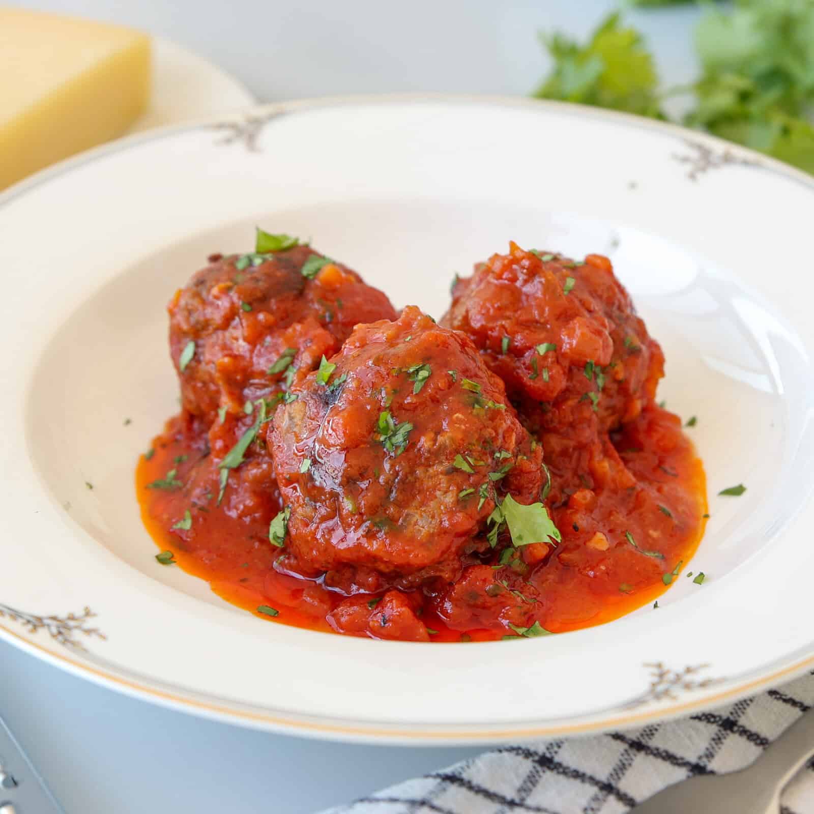 Three meatballs topped with tomato sauce and chopped parsley are served on a white plate. A block of cheese and fresh herbs are blurred in the background, along with part of a striped napkin.
