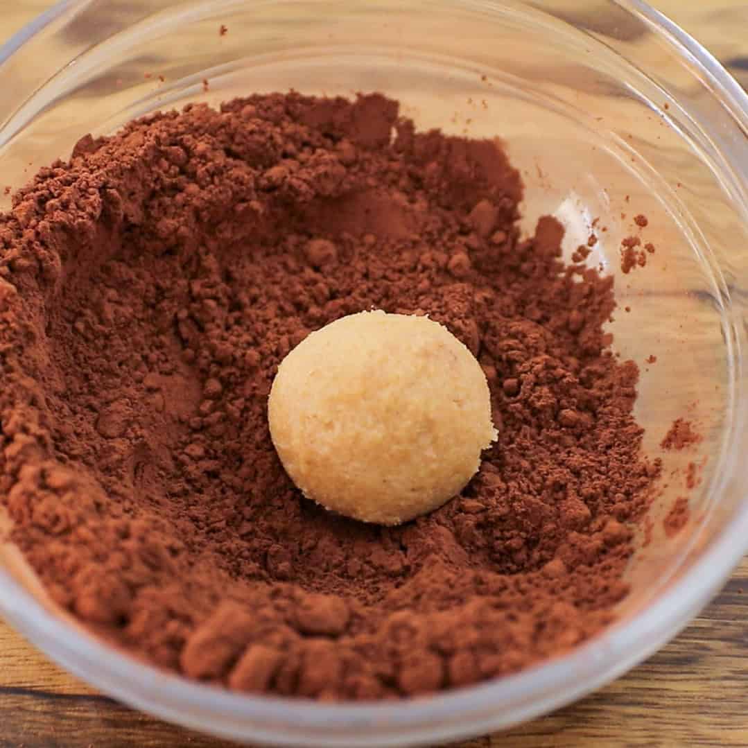 A round dough ball sits in a clear bowl filled with cocoa powder, ready to be coated. The bowl rests on a wooden surface.