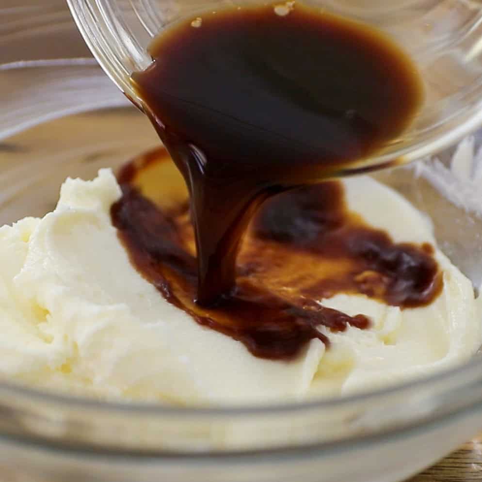 A glass bowl with creamy white mixture being topped by dark brown liquid poured from another bowl, likely a baking or dessert preparation step.
