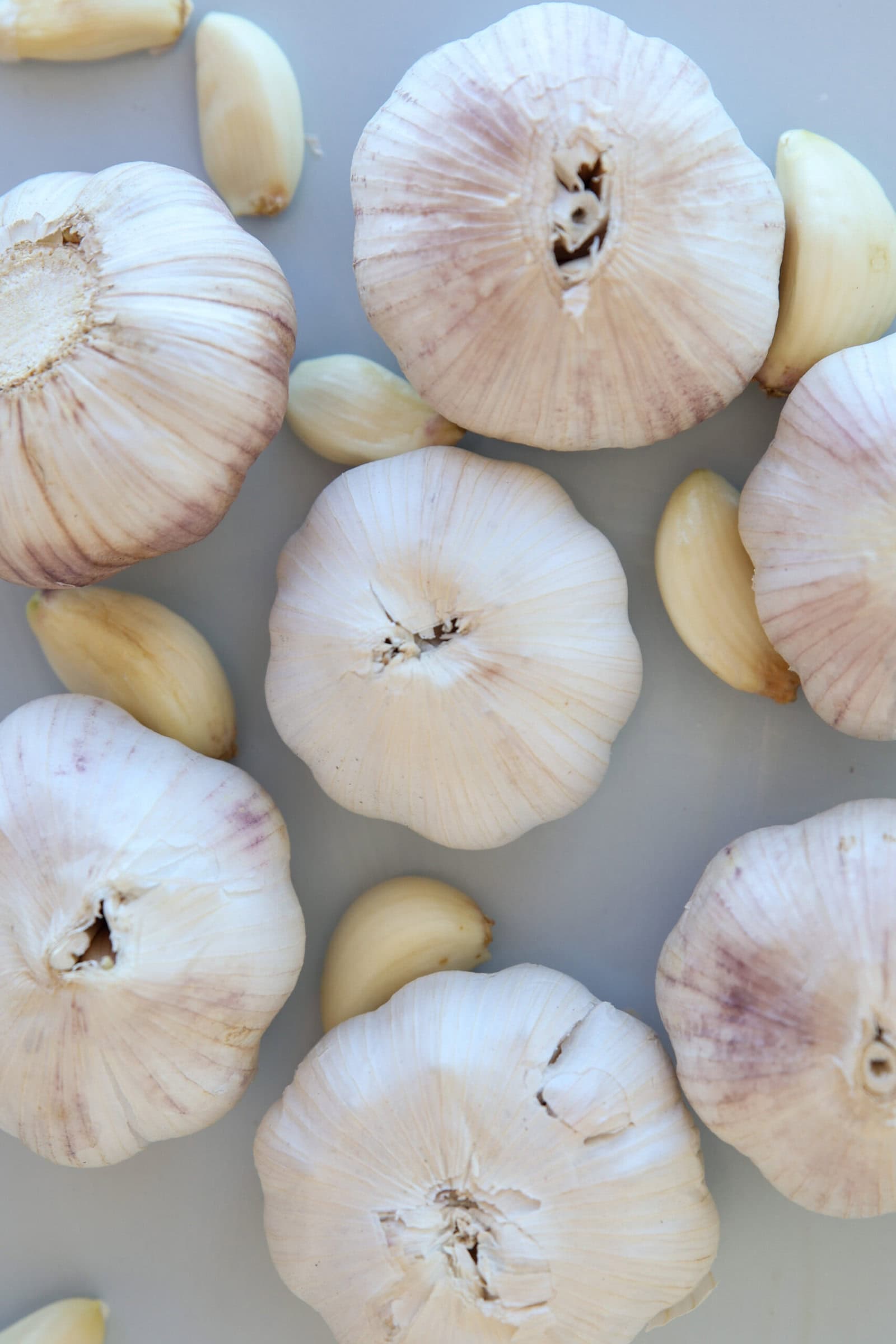 Whole garlic bulbs and several loose garlic cloves are arranged on a light-colored surface, viewed from above. The bulbs have a pale beige and purple tint, while the cloves are creamy white.