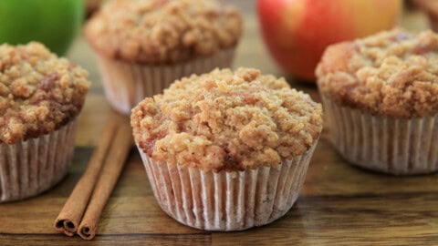 Easy Apple Crumble Muffins Recipe Fourapple crumble muffins are arranged on a wooden surface, with two cinnamon sticks and a red apple in the background. The muffins have a golden brown, crumbly topping.