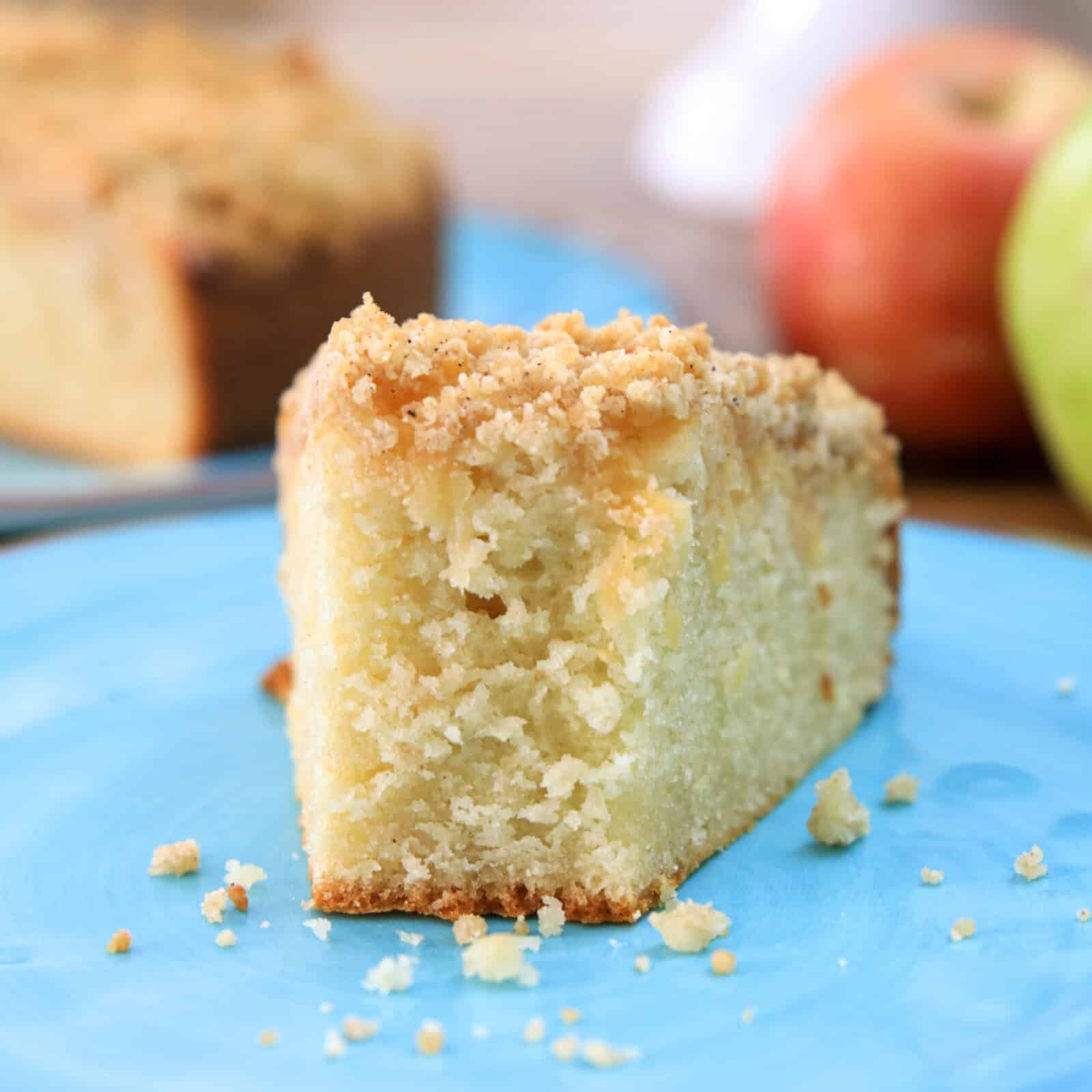 A close-up of a slice of crumb-topped coffee cake on a blue plate, with crumbs scattered around and blurred apples in the background.