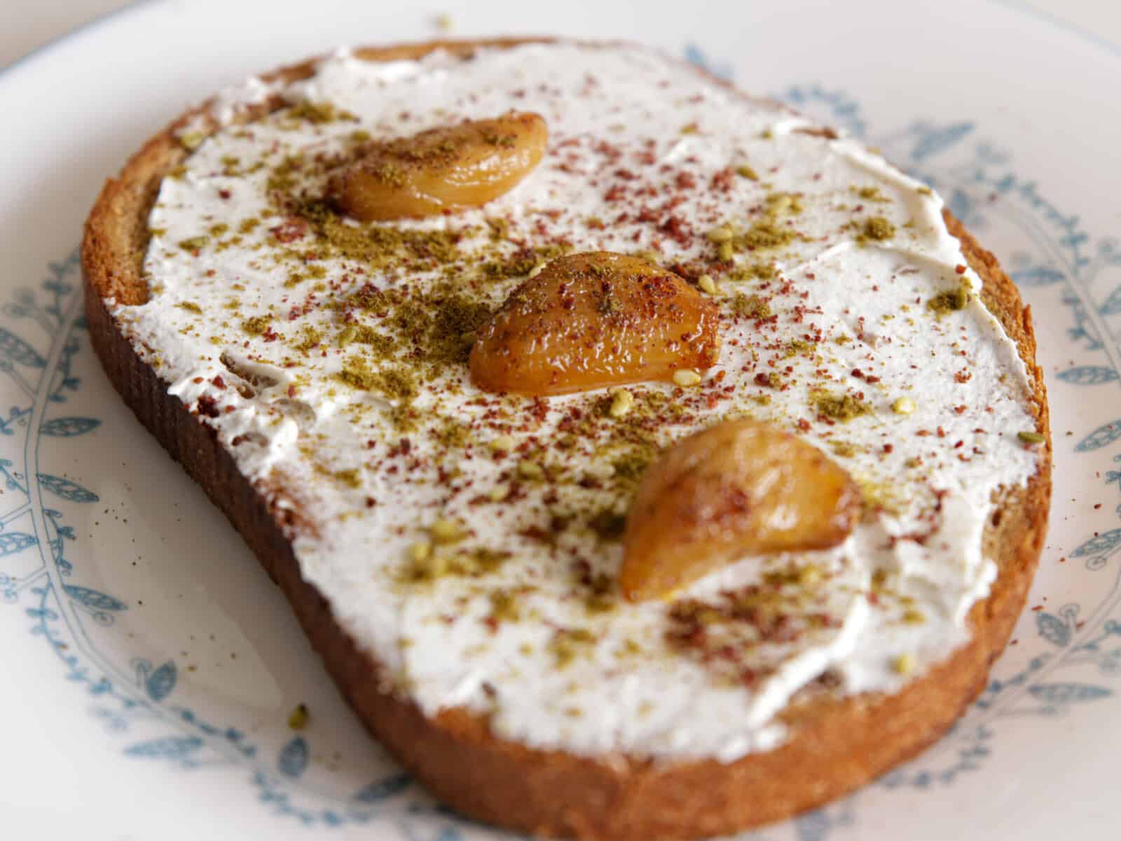 A slice of toast topped with a creamy white spread, roasted garlic cloves, and sprinkled spices, served on a white plate with a blue floral pattern.