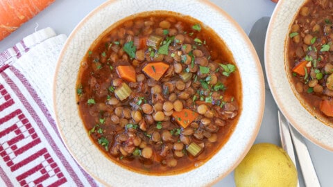 Easy Vegan Lentil Soup with Vegetables & Herbs A bowl of lentil soup with diced carrots, celery, and fresh herbs, served on a light plate. A striped towel, part of a lemon, and a spoon are visible beside the bowl.