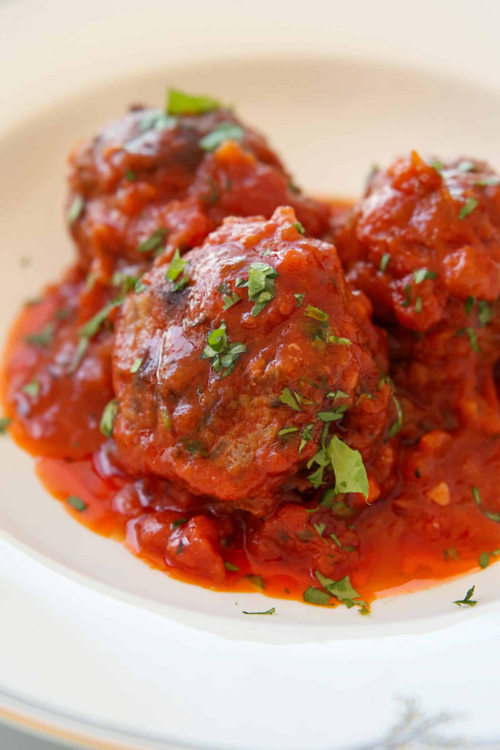 Three meatballs covered in tomato sauce and garnished with chopped fresh herbs are served on a white plate.