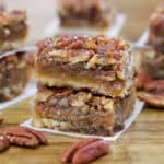 Two stacked pecan pie bars with a nutty topping and gooey caramel layer, resting on parchment paper. Whole pecans are scattered around on a wooden surface, with more bars in the blurred background.