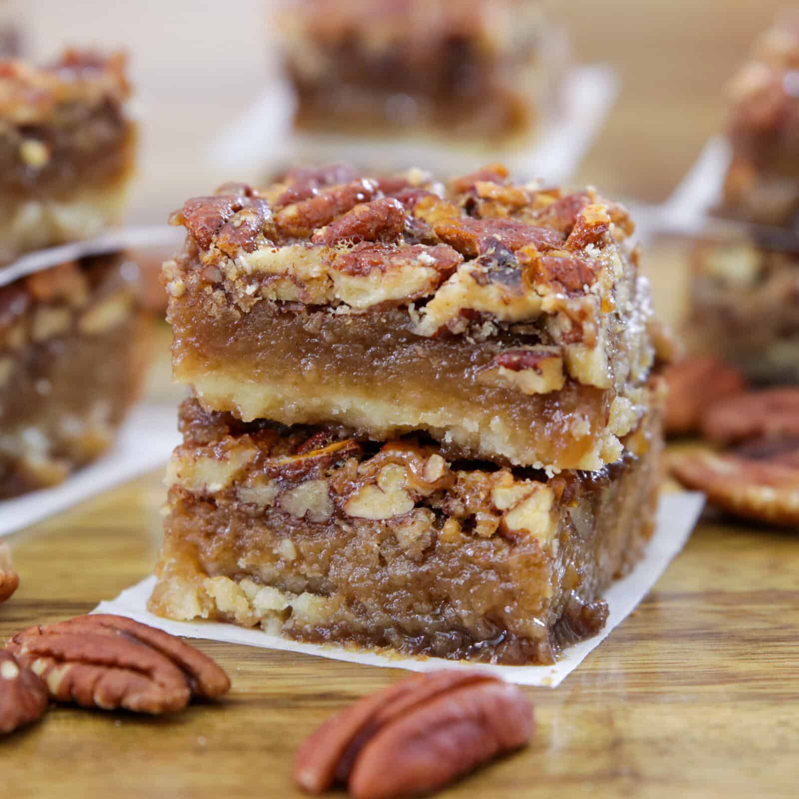 Two stacked pecan pie bars with a nutty topping and gooey caramel layer, resting on parchment paper. Whole pecans are scattered around on a wooden surface, with more bars in the blurred background.