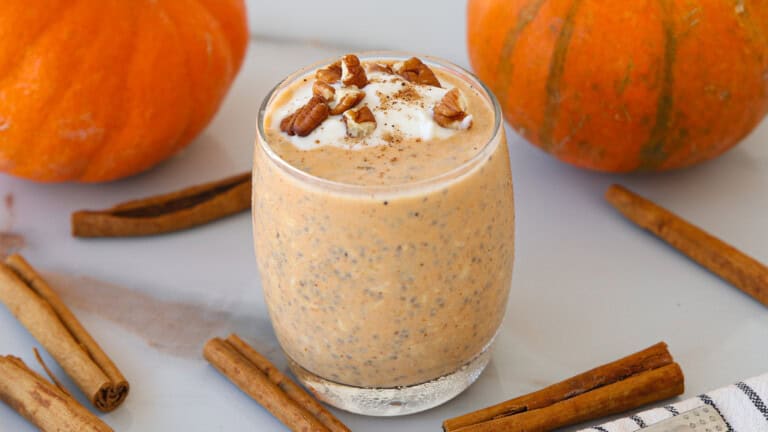 A glass of pumpkin spice overnight oats topped with whipped cream and chopped pecans sits on a light surface, surrounded by whole pumpkins and cinnamon sticks.