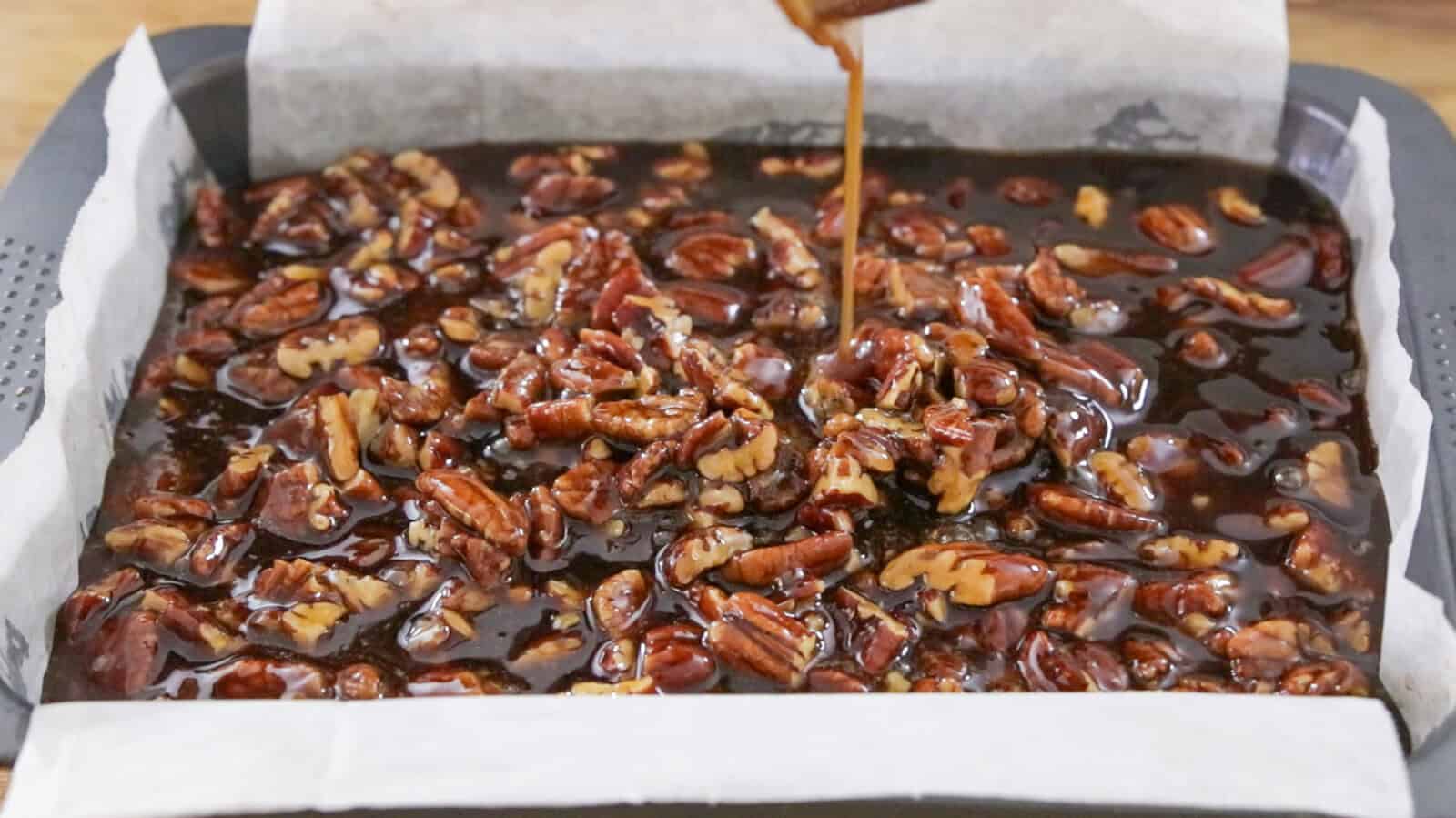 A parchment-lined baking tray filled with pecans and walnuts being covered in glossy caramel sauce, ready for baking or setting into a dessert.