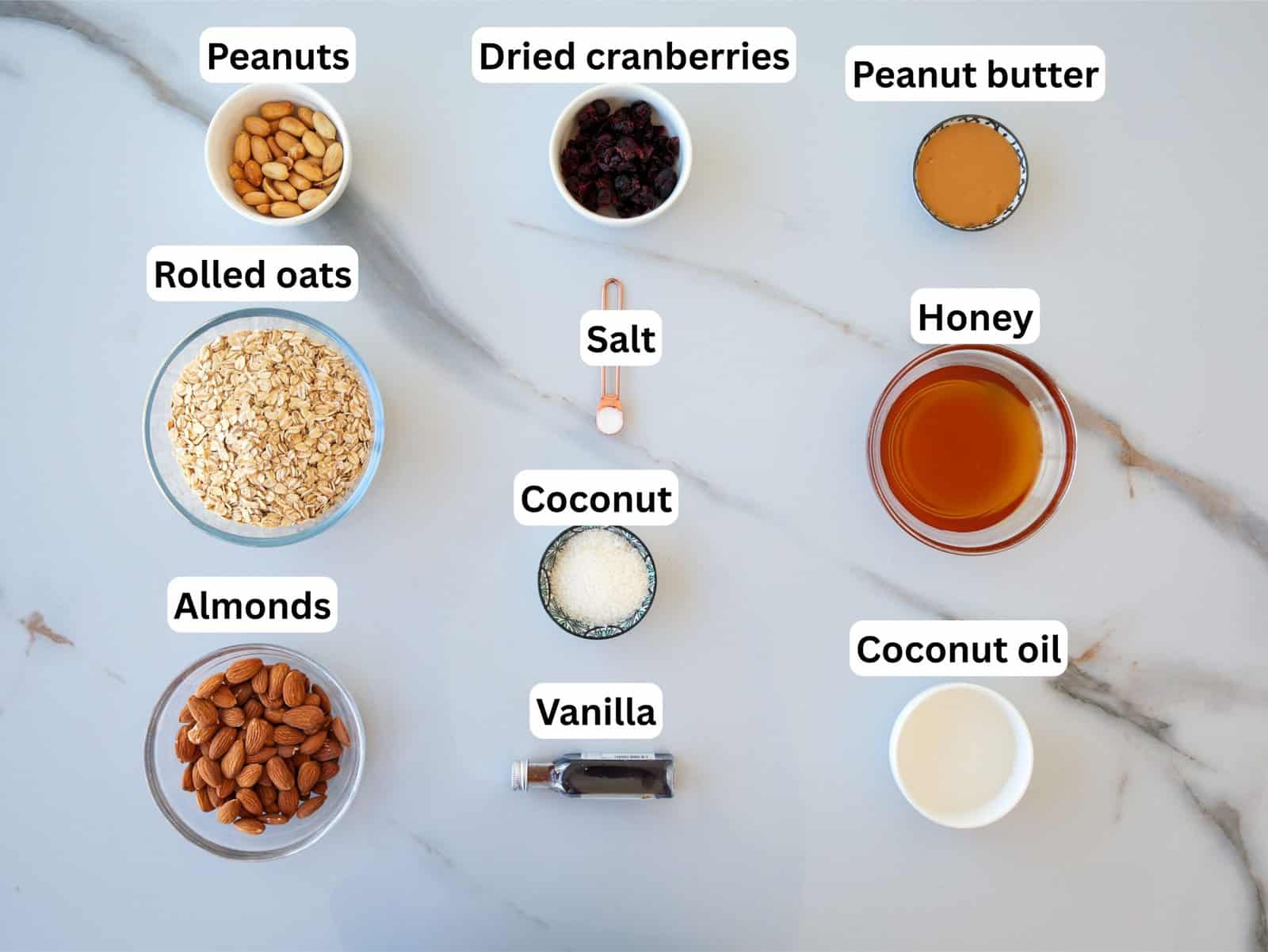 A flat lay of labeled ingredients on a marble surface: peanuts, rolled oats, almonds, dried cranberries, salt, coconut, vanilla extract, peanut butter, honey, and coconut oil in separate bowls and containers.