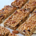 Close-up of several homemade granola bars with oats, nuts, and dried fruit, arranged in rows on parchment paper. The bars are golden brown and textured, with visible almonds and oats.