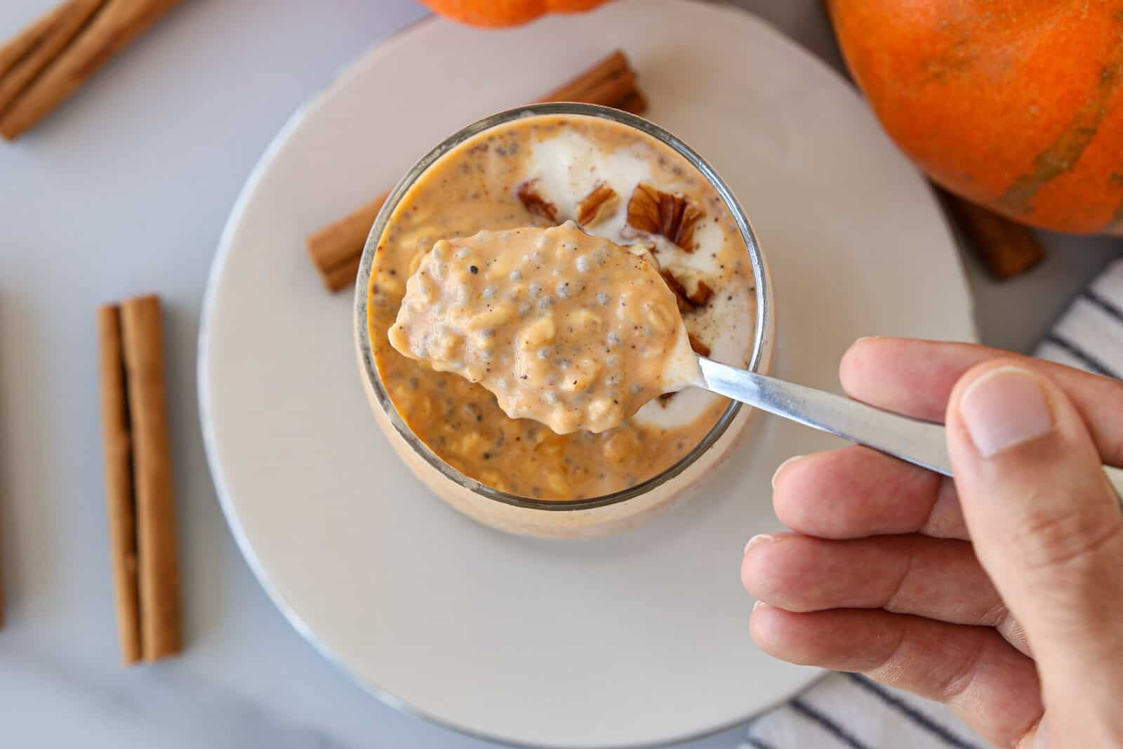 A hand holds a spoonful of pumpkin overnight oats  over a glass filled with the pudding, garnished with pecans. Cinnamon sticks and a pumpkin are nearby on a white plate and marble surface.